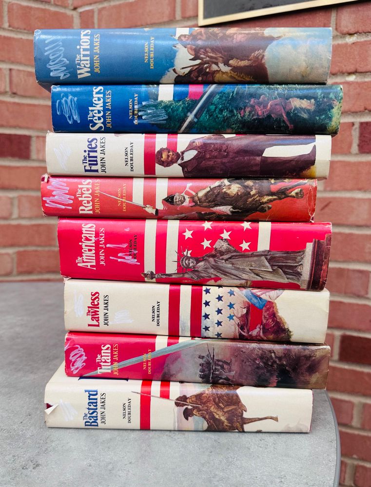 A stack of eight hardcover books stack one on top of another. They are the Kent Family Chronicles series by John James and are red white and blue in color.