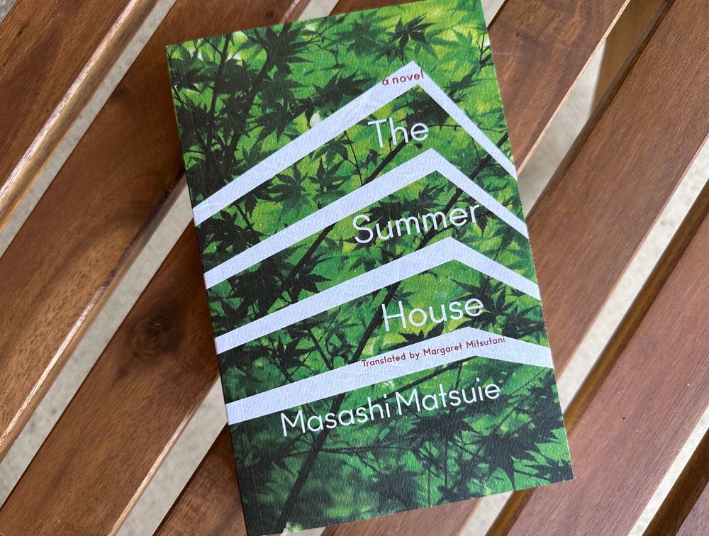 A book, The Summer House, written by Masashi Matsuie and translated by Margaret Mitsutani, sits atop brown wooden slats. The book itself is green with white striping.