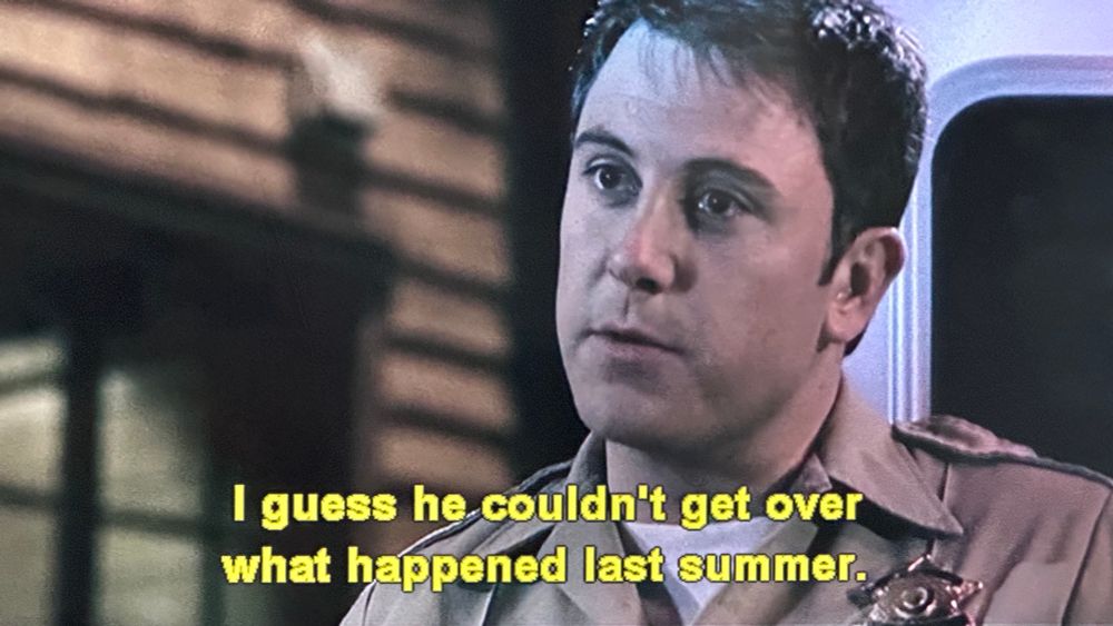 A sheriff stands in front of an ambulance, looking offscreen. Yellow subtitles beneath him read: 'I guess he couldn't get over what happened last summer.'