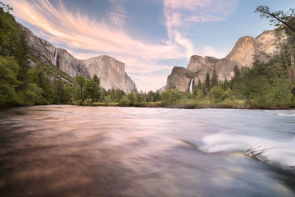 An overflowing Merced River courses through Yosemite Valley at sunset, with a very full Ribbon Fall (left) and Bridalveil Fall (right) flanking El Capitan.
