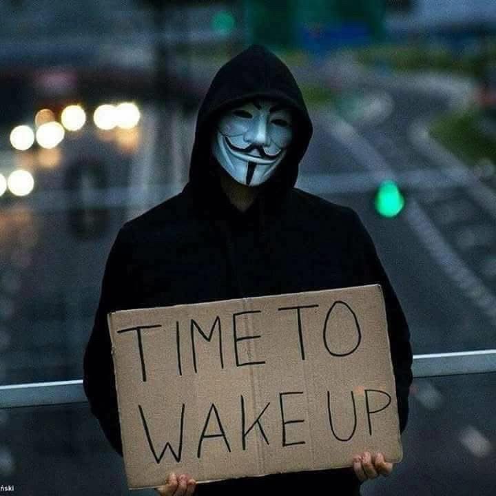 A man in an anonymous mask holds a sign that says,Time To Wake Up