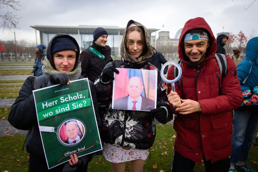 3 Personen mit Bundestag im Hintergrund: Links mit Plakat "Herr Scholz, wo sind Sie?", Mitte mit Foto von Olaf Scholz mit ausweichendem Blick, rechts mit einer Papplupe