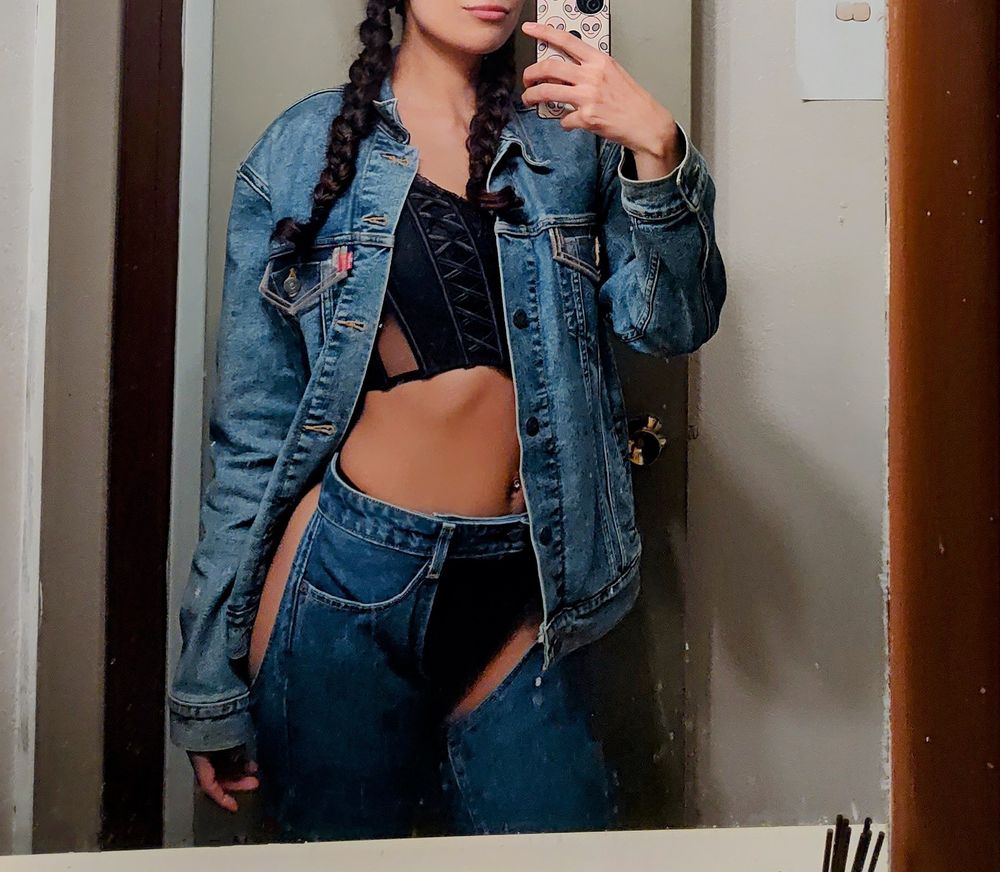 (Front view) A mirror selfie of Domina Machina wearing a denim jacket, denim chaps, a black corset top, and a black thong. Their hair is in 2 braids
