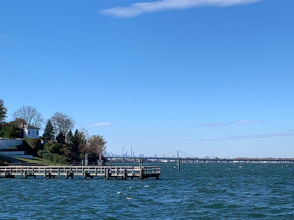 Blue water with a house, trees, and a dock at left, big blue sky with bridge and city skyline in the far distance. 