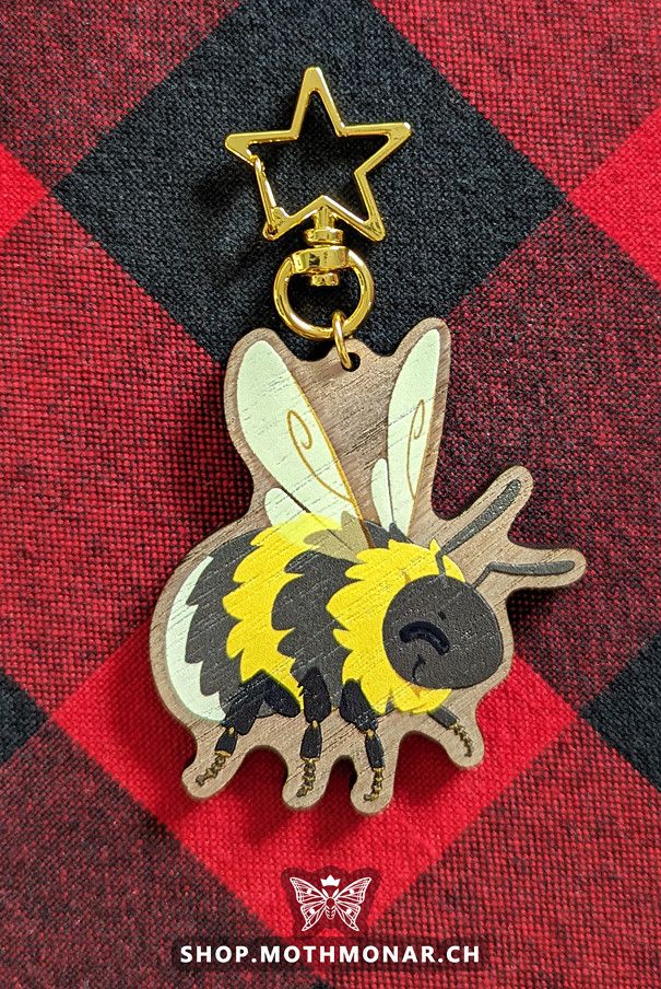 Photo of a wooden printed bumblebee charm on a red plaid cloth. The cartoon bee is facing right with a smiling expression. Top of the charm has a star clasp.
