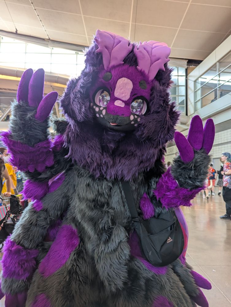 Cute black and purple moth fursuiter with lavender antennae, pink freckles, and large shiny purple eyes waving with two hands at the viewer. They have a black messenger bag to one side. Behind them a bit of a convention hall and a few people are visible.