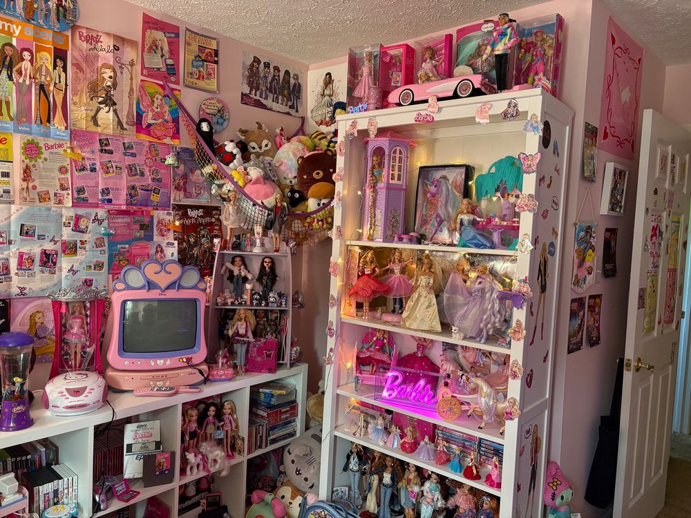 Office filled with Y2K nostalgia featuring Disney Princess CRT, Hello Kitty radio/CD player, stuffed animals, plug and plays, Barbies, Bratz, and My Scene dolls, and posters/advertisements on the wall. 