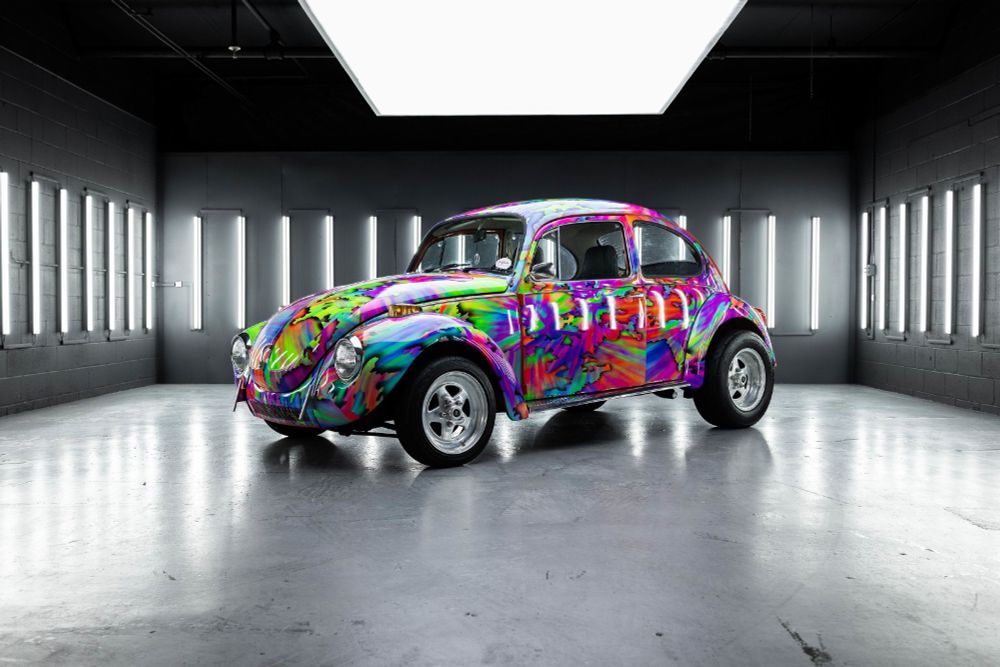 A 1972 VW Beatle in a psychedelic rainbow wrap. The rear tires are much larger than the front. It's being shown off in a well-lit garage.
