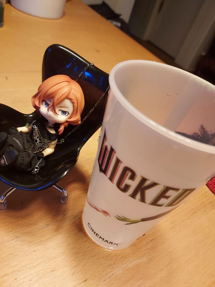 Chuuya Nenderoid doll in black chair next to a souvenir cup from the movie Wicked 