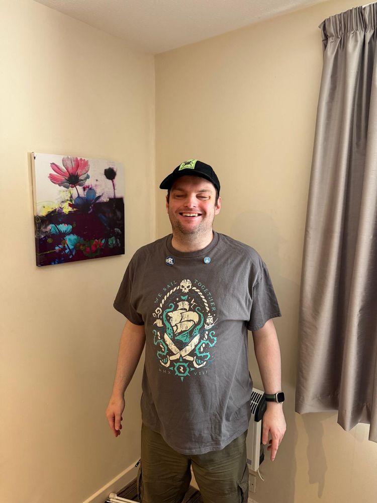 Sightless stands smiling, wearing his trademark cap with his logo on it and a shirt with “We shall sail together” on the front. Two pins, one on each side are visible.