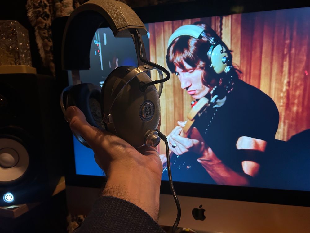Photo of a computer screen displaying Pink Floyd's Live at Pompeii, with Roger Waters recording his bass part for Us and Them, wearing headphones that look identical to the pair of headphones that i am holding up next to the screen in comparison