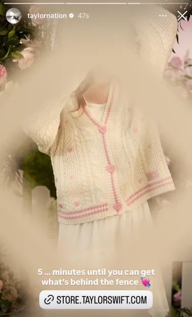 A screenshot from Taylor Nation of the new lover cardigan. Similar to the OG but with baby pink lines and pink hearts. The buttons are also pink. The image is of a model showing off the cardigan but the camera is pushing through what appears to be a lattice fence similar to the five holes in the fence post from pre lover
