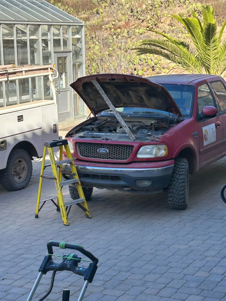 A red pickup truck with the hood held open by a long 2x4