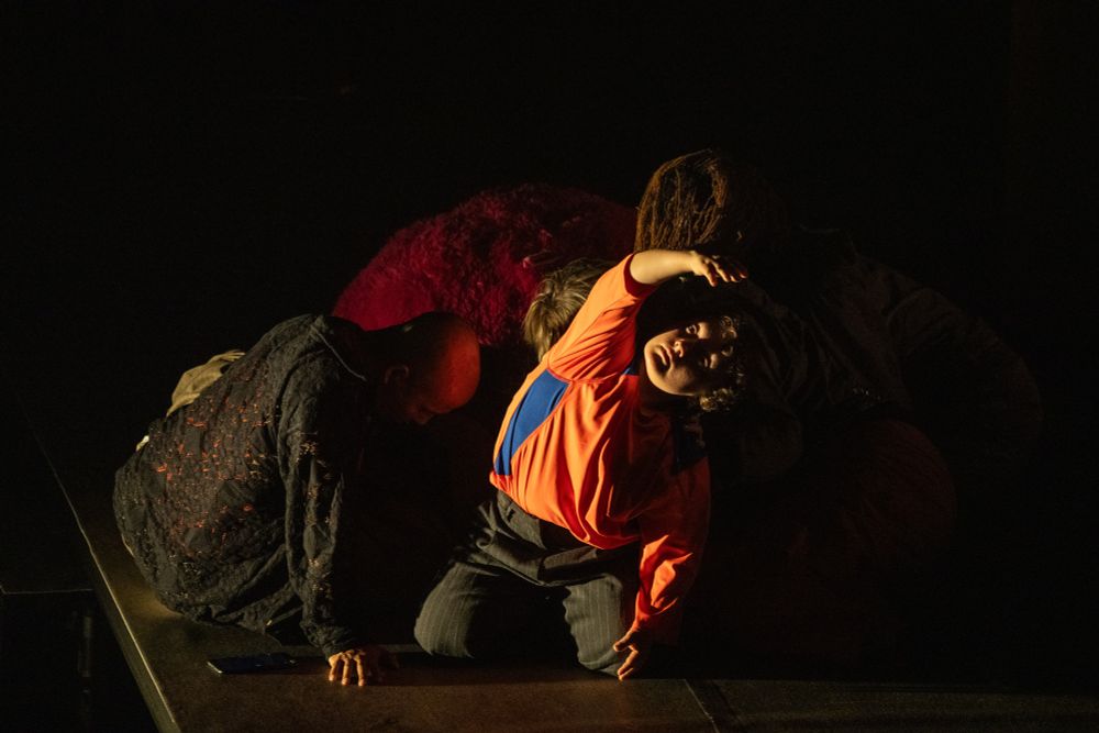 Close-up of a dancer under a spotlight, captured mid-performance while leaning to one side with one hand raised. In the background, a group of dancers move closely together in soft focus, adding depth and energy to the stage scene.