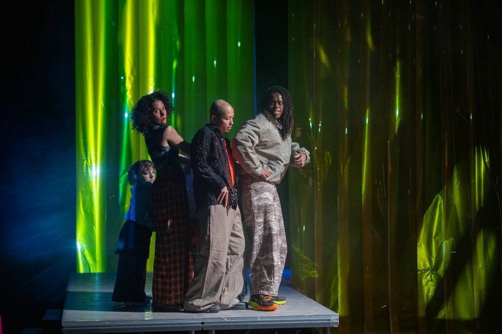 A group of dancers performing in a line on a stage platform. The background features vibrant green projections, casting dynamic patterns that enhance the visual energy of the performance.