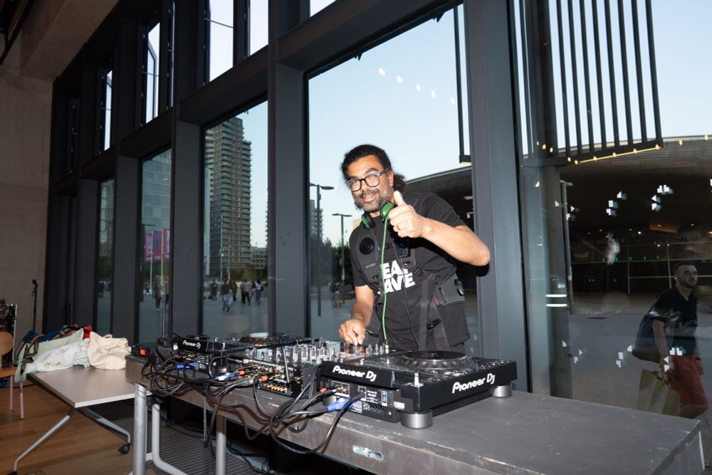 A DJ stands behind a DJ station in front of a large glass window, smiling and giving a thumbs-up toward the camera.