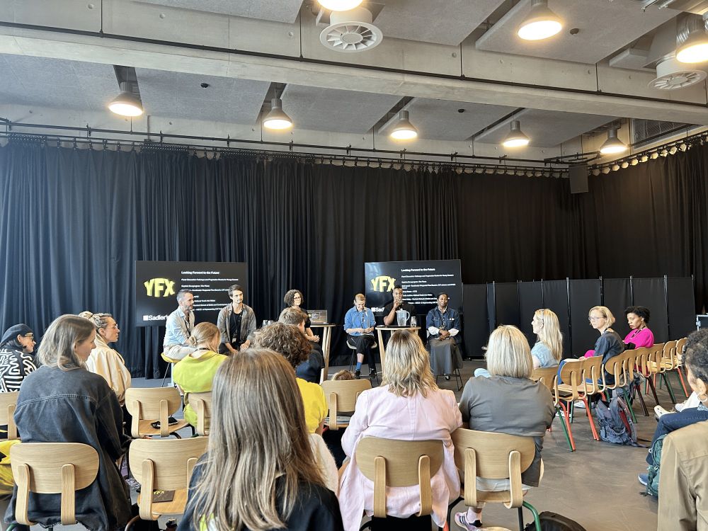 Six panelists seated on stage engage in a discussion in front of a live audience, whose backs are facing the camera. Two large screens behind the panel display a presentation featuring Sadler's Wells East's festival logo ‘YFX.’