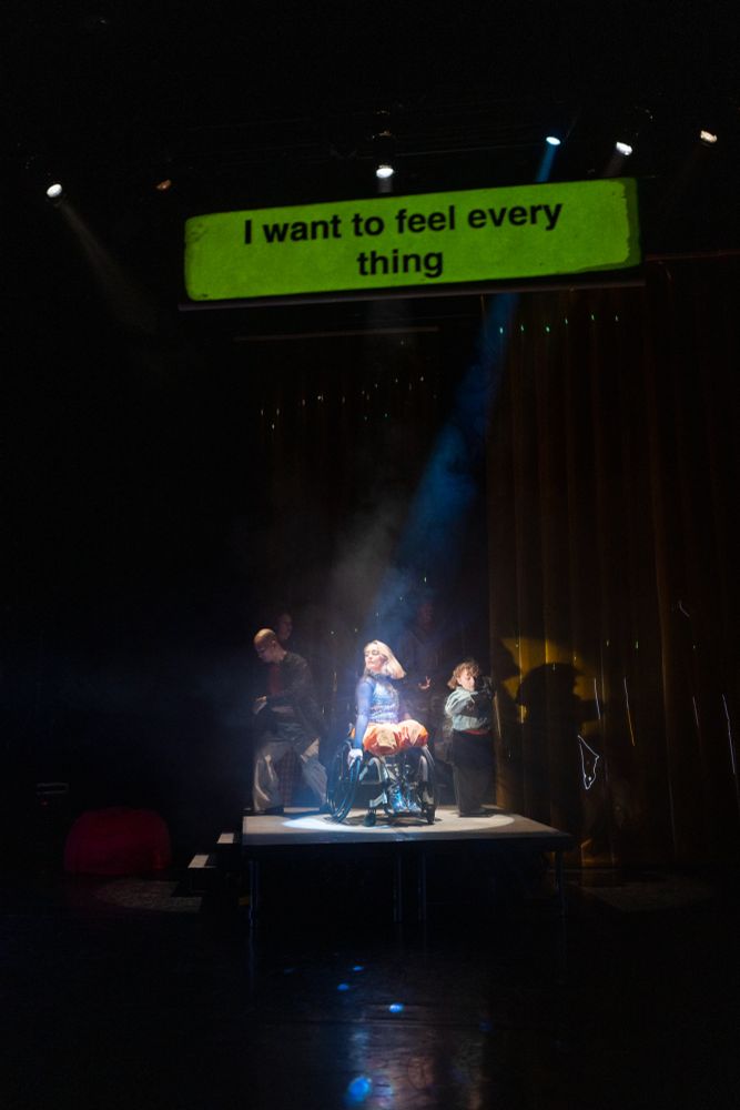 A group of performers on stage, with three dancers in the spotlight at the front, captured mid-movement. Above them, a green caption screen reads: “I want to feel everything,” highlighting the emotional intensity of the scene.