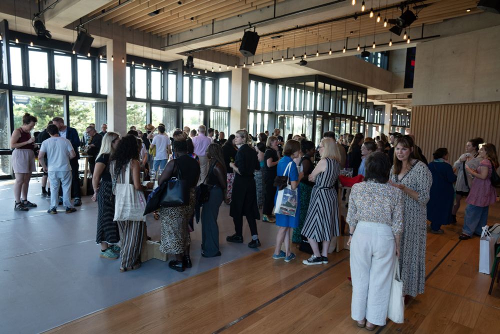 A large group of people, gathered in smaller clusters, chat and enjoy drinks in a spacious indoor venue.