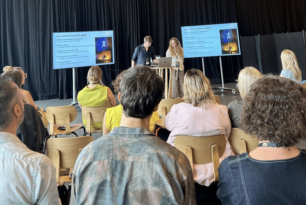 Two Candoco staff members give a live presentation on the inclusive practices developed during the creation of Candoco’s latest work, Over and Over (and over again). They stand in front of an audience, whose backs face the camera. Behind the presenters, two large screens display slides with text and images from the production.