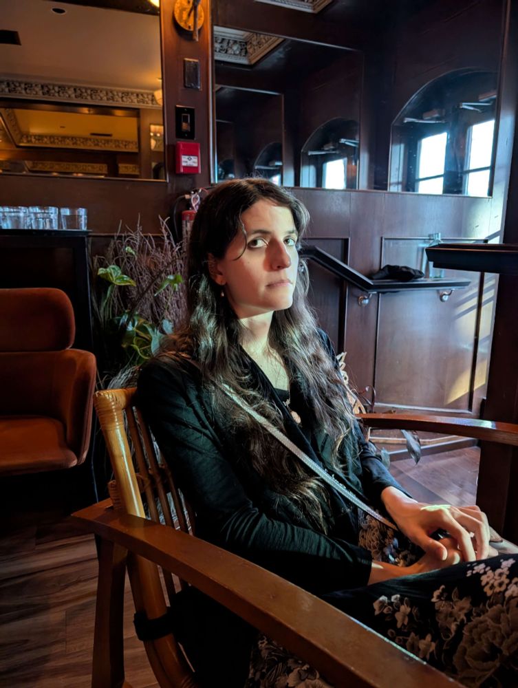 A long-haired woman in a monochrome floral dress and a black jacket sits in a wooden chair. She looks at the camera with an aloof expression while the light streams in onto her through the window.