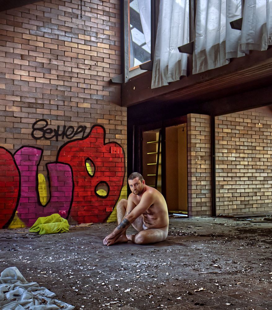 A nude masc appearing model poses sitting in an exposed abandoned house. 