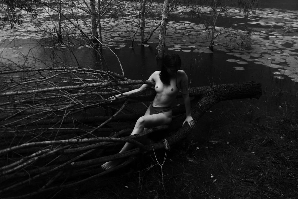 black and white photograph of a topless model sitting amongst a recently fallen tree on the edge of a lake. The light is washing across his body while their face is covered in a shadow.