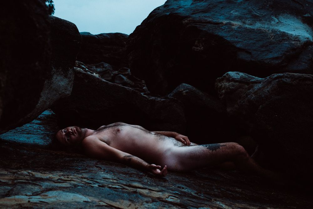 A nude AMAB model lays among large rocks on a rainy day 