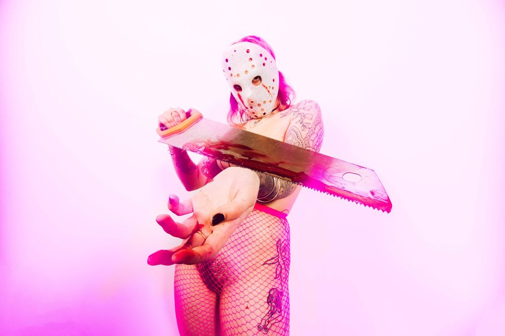 A nude model stands holding a bloody saw to their wrist wearing pink fishnets and a bloody hockey mask