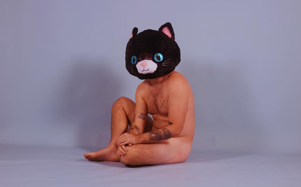 A photo of a nude model wearing a large cat head sitting against a studio backdrop.