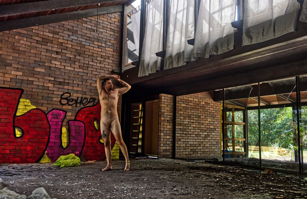 A nude masc appearing model poses standing with their arms above their head in an exposed abandoned house. 