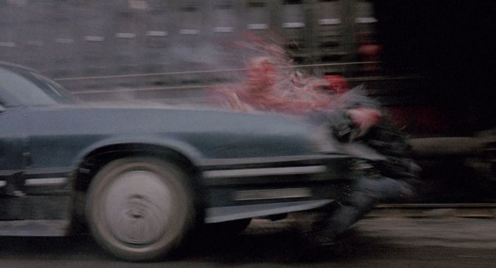 A scene from the sci-fi action film RoboCop, wherein a man who has been exposed to toxic chemicals is hit by a car, which causes him to disintegrate like a mummy made of pork trimmings wrapped in wet toilet paper. His face is visibly detaching from his body and hurtling towards the windshield. The car in question is the 6000 SUX, advertised in the film as “big”, with an MPG of about eight. From a production standpoint, it’s a ’76 Oldsmobile built up with a fiberglass exterior. This scene is arguably more iconic and widely remembered than the ultimate villain’s defeat at the end of the film, although the man being dispatched here is a relatively minor antagonist. This is largely due to the quality and spectacular nature of the practical effects in play.