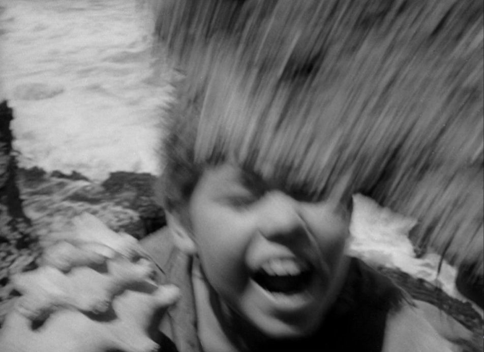 A scene from the adventure film Lord of the Flies. A young boy stands on some rocks at the edge of a churning sea. He is very close to the camera. In one hand he holds a conch shell. His mouth is open and eyes closed in surprise. An enormous rock falls into frame. The whole image blurs with the violence of the action.