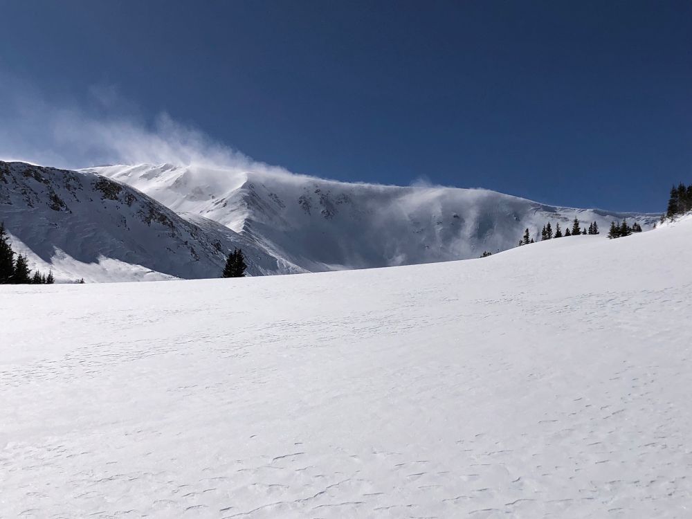 Mountain ridge in the background with snow blowing off to the top left. Foreground is an expanse of smooth snow. You can see a few evergreen trees in the distance.