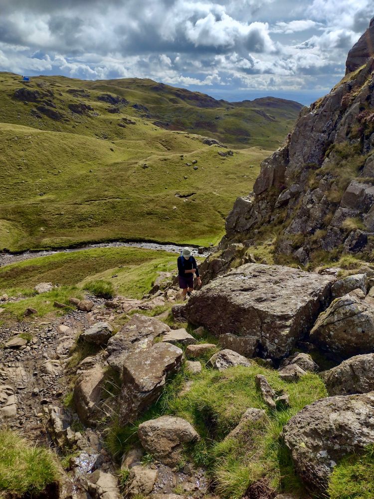 an old man struggles wearily up the path behind Pavey Ark, Gt Langdale, August 2023. it's all very green, with boulders in the foreground, the lower shoulder of the crag to the right. a stream cuts the middle ground. dirty yet luminous cloud in the distance