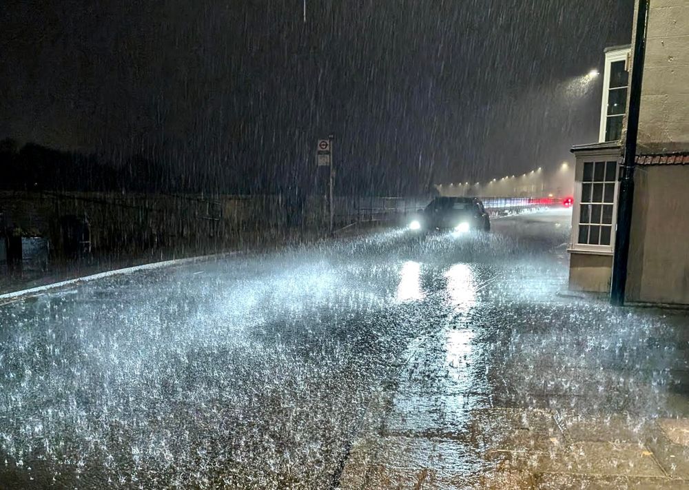 Night from under Barns Bridge, pouring rain full of light, roaf full of reflections, a car approaching fast from the direction of the Bull's Head. someone says: "More water on the road than in the river."