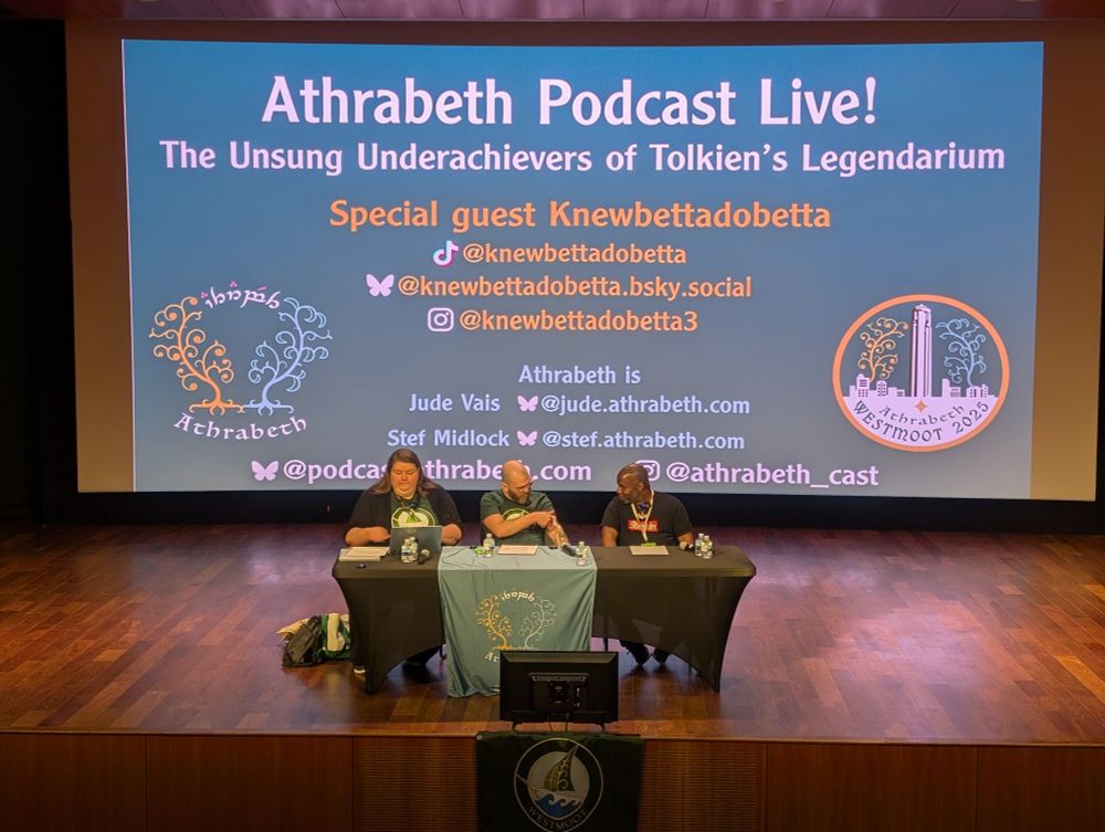 three people sitting on a stage behind a table with a slide deck projected behind them that reads Athrabeth Podcast Live!