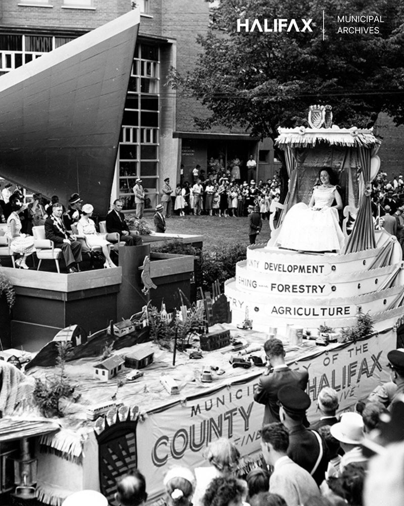 A large parade float with a banner that says, 'Municipality of the County of Halifax' and another that says 'Development, Forestry, Agriculture'. There are miniature buildings, vehicles and trees on the flatbed of the float. A young woman wearing a light-coloured gown sits on a platform at the back of the float. The crowd watches on from the sidelines - some people are seated on a platform.