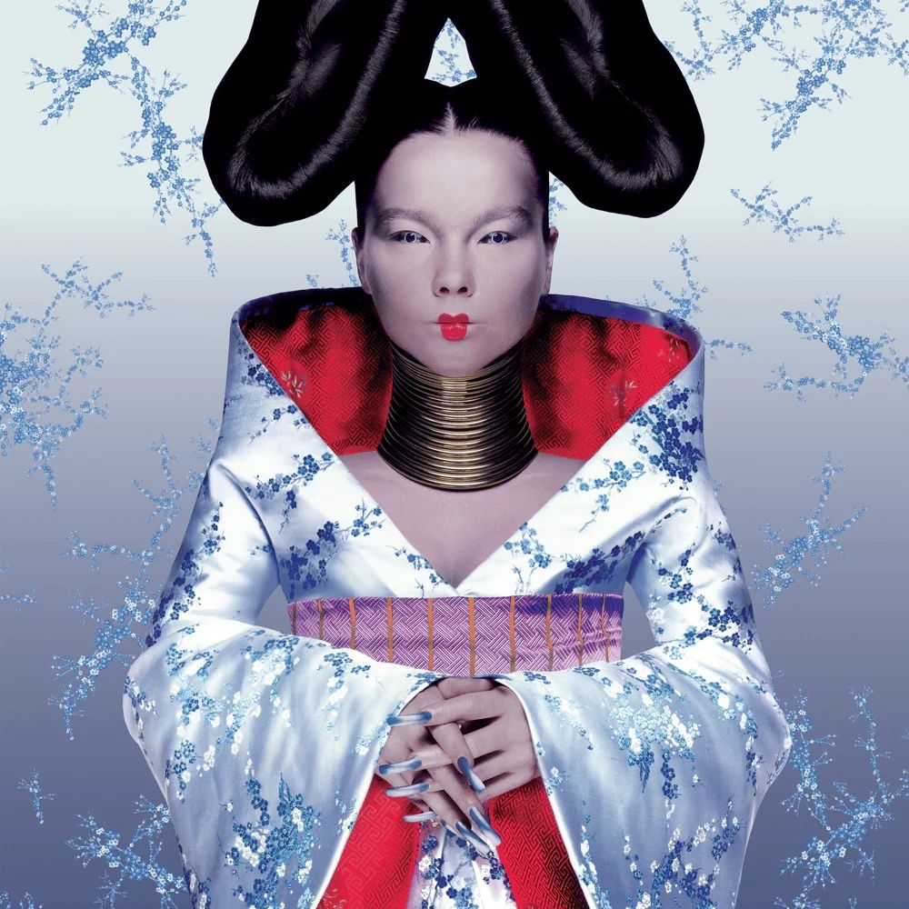 Cover of album “Homogenic” by Icelandic singer Björk featuring her, a woman in her thirties, in front of a grey background wearing a silver kimono, red on the inside, long nails, a coiled neckpiece and geisha-like makeup.