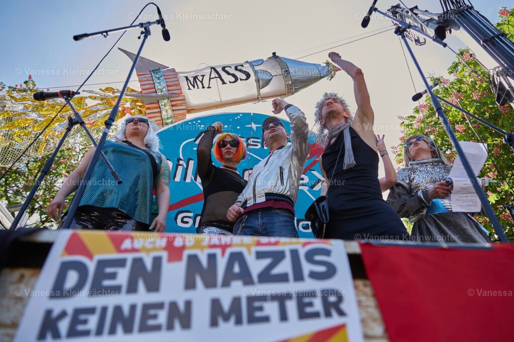 Der Rattenchor, fünf queere Menschen, teilweise mit erhobenen Fäusten, auf einer Bühne. Um sie herum Mikrofone, im Hintergrund die MyASS-Rakete, unscharf im Vordergrund die Worte "Den Nazis keinen Meter".