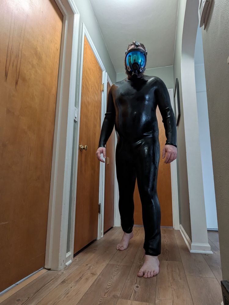 Person in silicone catsuit and mirrored gas mask steps toward and looks directly at the camera. In the visor you can see there's a mirror in which you can see the person again. The shot is from low and the backdrop is a hallway with 5 doors, one open. 