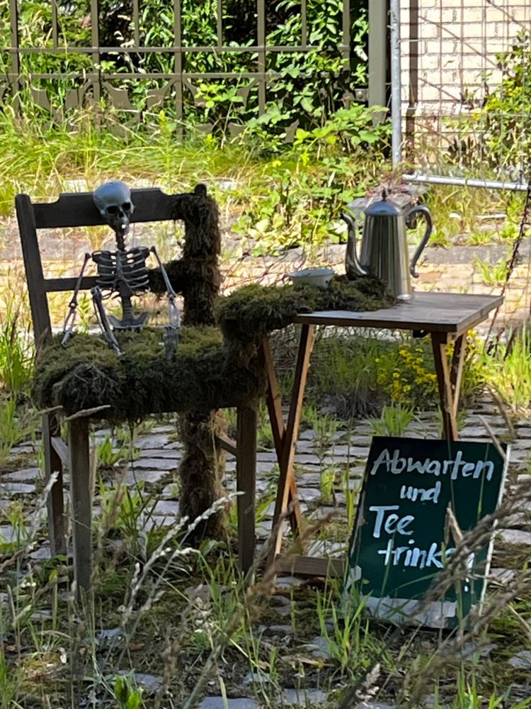 Ein kleiner Tisch mit Teekanne und Tasse, daneben ein vermooster Stuhl auf welchem ein Skelett sitzt. An den Tisch ist eine Tafel gelehnt mit der Aufschrift: Abwarten und Tee trinken. 