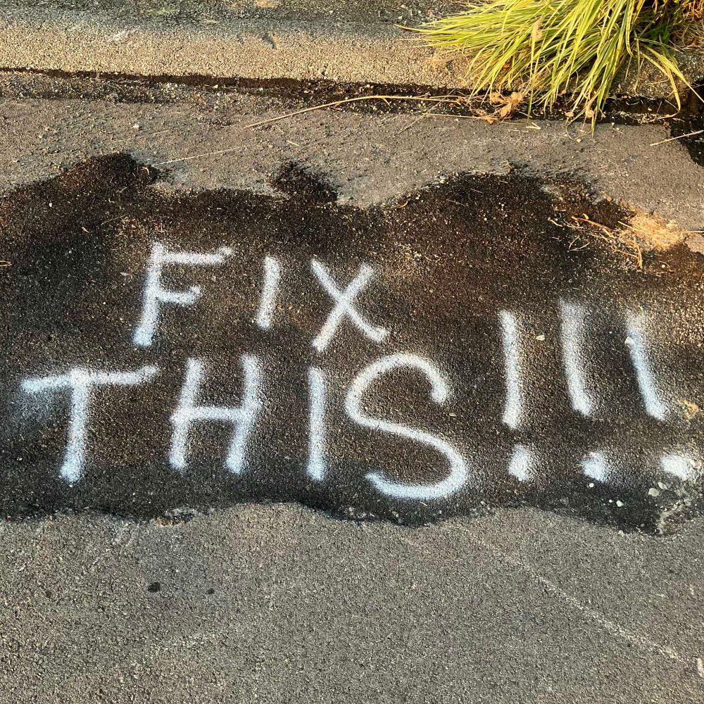 A message sprayed on an asphalt patch on a street, saying “FIX THIS!!!”