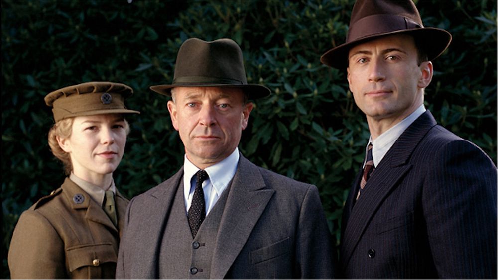 An upper body and headshot of three titular characters from the British TV show “Foyle’s War”. On the left is the female driver Sam Stuart in uniform and hat of the MTC, in the centre is protagonist DCS Foyle in a suit and fedora, and on the right is Sgt. Milner in a suit and fedora.
