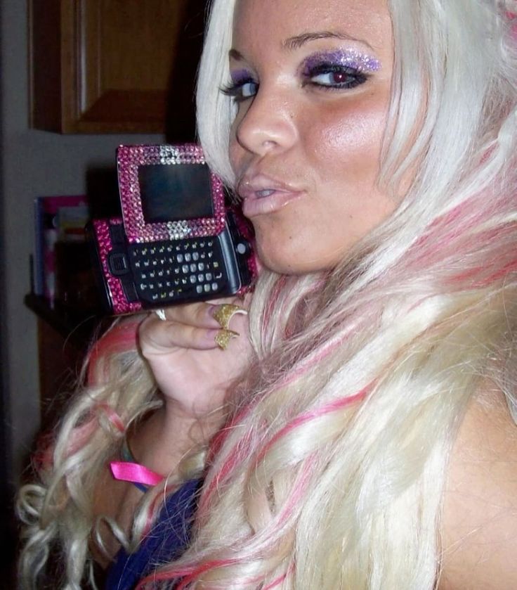 The image shows a young Trisha Paytas during the 2000s posing with a Sidekick phone bedazzled in pink and silver gems held up to her face. She is pouting slightly. She has long, platinum blonde hair with pink streaks styled in loose curls cascading over her shoulders. She’s wearing purple glittery eyeshadow, thick black eyeliner, bronzed cheeks, and glossy, lined lips in a nude shade. She has a gold glittery nail extensions in a “duck” tip shape.