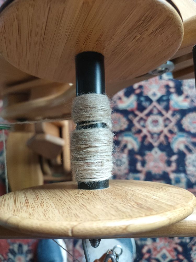 A spinning wheel bobbin with a little bit of fine spinning: NZ Halfbred wool with 30% tussah silk. The spinning wheel is sitting on an eastern pile carpet with a geometric design of stylised birds and flowers.