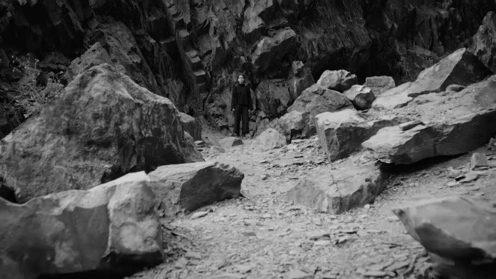 A woman in black clothing stands amid huge broken rocks in front of a jagged cliff face.