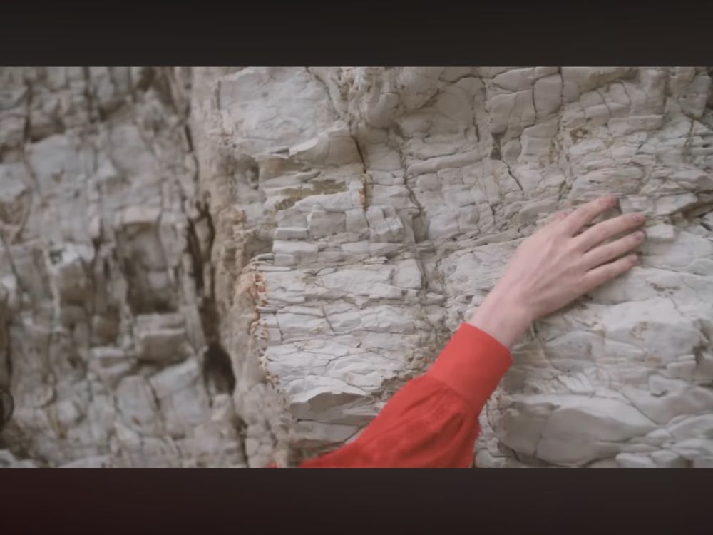 Color image of Margaux Sauve running her hand along a vertical rock face. red sleeve, beige rock