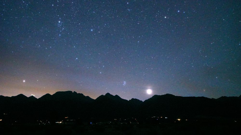 The sky is dark, Venus rises over the silhouette of the Rocky Mountains, magnified by a thin layer of clouds. Pleiades is left and up a little from Venus. The sky is full of stars. The lights of Crestone and Baca Grande homes sprinkle the foreground. There is some light pollution visible on the left from the Front Range.