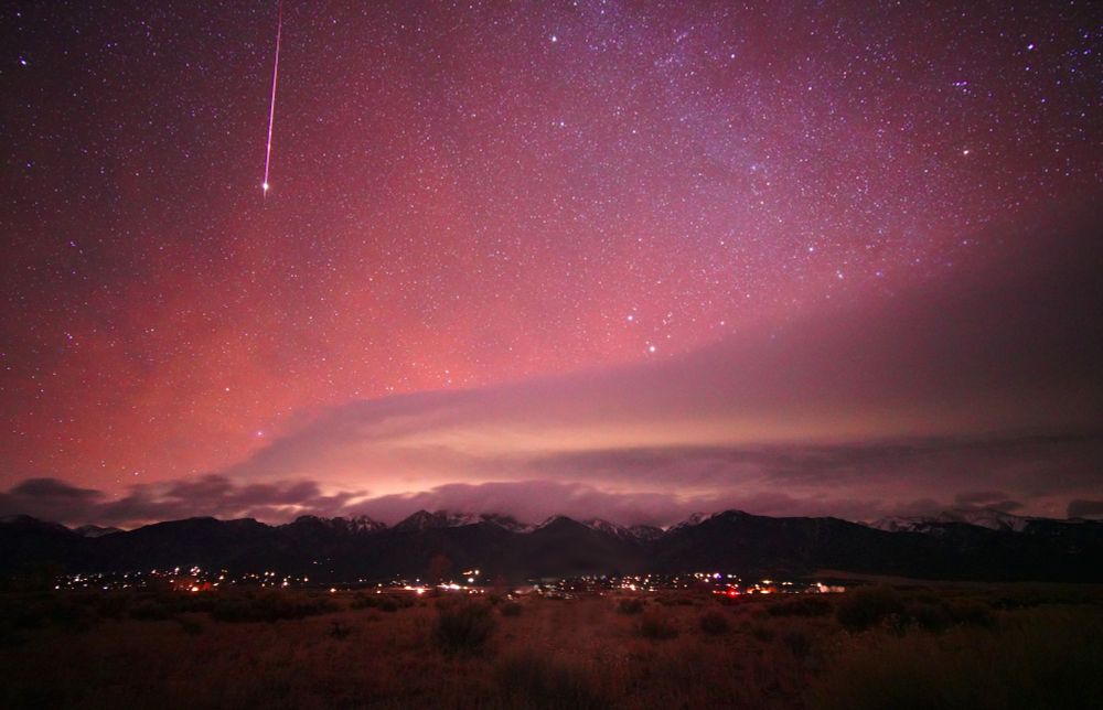 A brilliant pink-purple meteor streaks down top left, about 1/3rd of the frame. It has a terminal flash. The sky is dark maroon on the left merging to purple on the right, and orange below. There's an epic shelf cloud sitting over the mountains, reflecting light pollution from the Colorado Springs area. The Sangre de Cristo range of the Rocky Mountains are capped with snow and lower clouds. Many house lights of Crestone and Baca Grande are visible at the base of the mountains. The near foreground is dry grass and chamisa